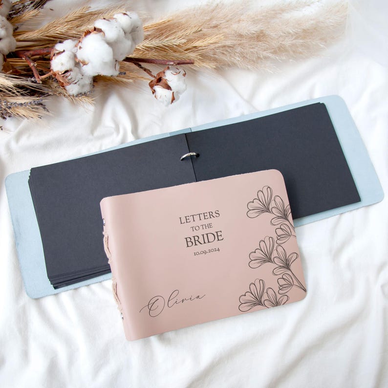 May include: A pink leather-bound journal with the text "Letters to the Bride" and the date "10.00.2024" engraved on the front cover. The journal has a black interior and a blue cover.