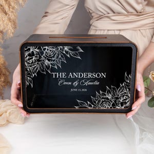 May include: A black wooden card box with a slot on top, featuring white floral designs and the text "THE ANDERSON Owen & Amelia JUNE 13, 2026". The box has a dark wood frame and is held by a person.