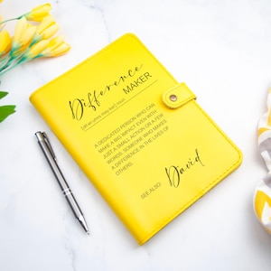 Könnte beinhalten: Ein gelbes, ledergebundenes Notizbuch mit dem Wort "Difference Maker", definiert als "eine engagierte Person, die selbst mit einer kleinen Handlung oder wenigen Worten einen großen Einfluss haben kann. Jemand, der das Leben anderer Menschen verändert." Der Name "David" steht unter der Definition.