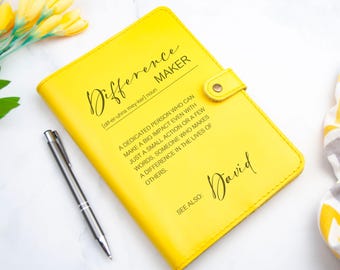 May include: A yellow leather-bound journal with the word "Difference Maker" defined as "a dedicated person who can make a big impact even with just a small action or a few words. Someone who makes a difference in the lives of others." The name "David" is written below the definition.