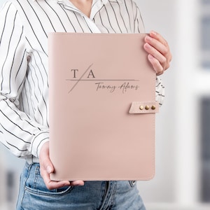 May include: A dusty rose colored leather-like portfolio folder featuring a horizontal line design with the initials T A and the name Tammy Adams printed in dark gray. The folder includes a side tab closure secured with three small metallic studs.