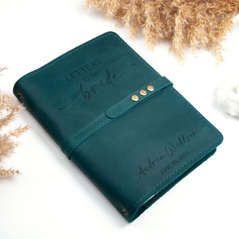 Op de afbeelding: Turkoois lederen dagboek met een band en gouden accenten. Het dagboek is gegraveerd met de tekst "Letters to the Bride" en "Audrie Willson June 15, 2024".