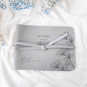 May include: Gray leather-bound journal with a floral design and the text "Letters to the Bride" and the name "Olivia" engraved on the front cover.