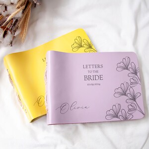 May include: Two leather-bound journals with floral designs. The purple journal has the text "Letters to the Bride 10.09.2024 Olivia" and a floral design. The yellow journal has a floral design.