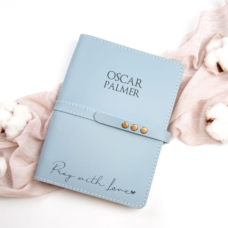 K&ouml;nnte beinhalten: Ein hellblaues Lederjournal mit dem Namen "Oscar Palmer" und dem Spruch "Pray with love" auf der Vorderseite. Das Journal hat einen goldenen Verschlussriemen mit drei goldenen Nieten.