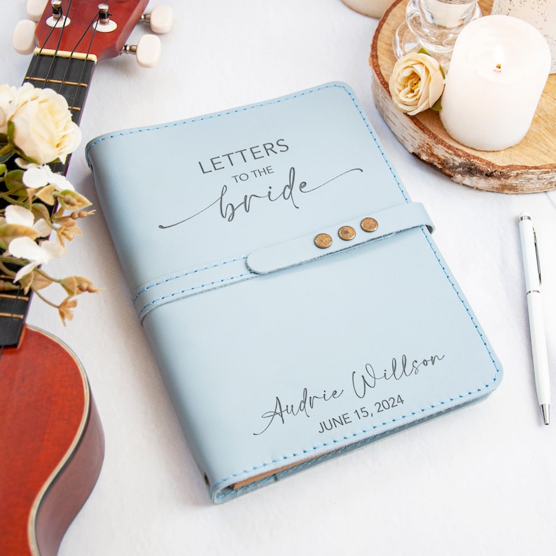 Op de afbeelding: Een lichtblauw lederen gebonden dagboek met de tekst "Letters to the bride" en een decoratief handschrift. Het dagboek heeft een bruine leren band met messing accenten en de naam "Audrie Willson" en de datum "June 15, 2024" zijn gegraveerd op de voorzijde.