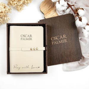K&ouml;nnte beinhalten: Ein cremefarbenes Lederjournal mit dem Namen "Oscar Palmer" auf dem vorderen Einband. Das Journal befindet sich in einer Holzkiste mit dem gleichen Namen auf dem Deckel. Das Journal hat die Worte "Pray with love" auf der Unterseite eingraviert.