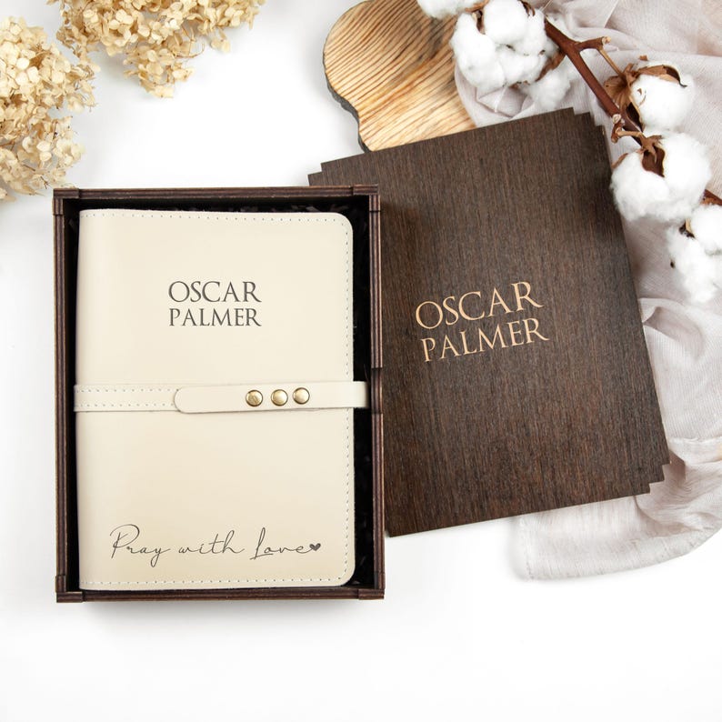 K&ouml;nnte beinhalten: Ein cremefarbenes Lederjournal mit dem Namen "Oscar Palmer" auf dem vorderen Einband. Das Journal befindet sich in einer Holzkiste mit dem gleichen Namen auf dem Deckel. Das Journal hat die Worte "Pray with love" auf der Unterseite eingraviert.