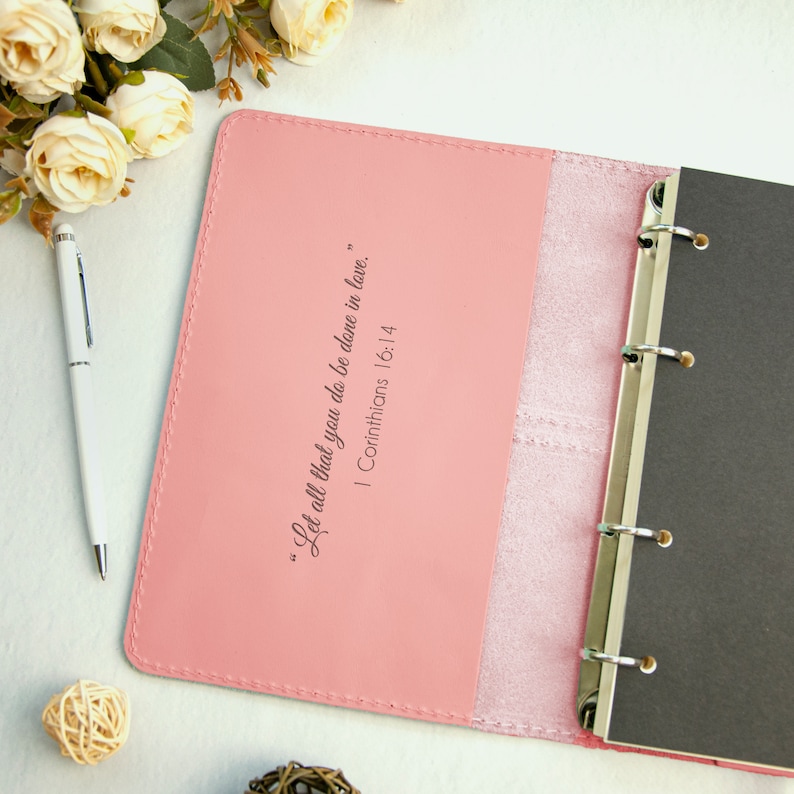 Op de afbeelding: Een roze lederen gebonden dagboek met een citaat uit de Bijbel, "Laat alles wat je doet, in liefde gedaan worden." 1 Korinti&euml;rs 16:14, gedrukt op de voorzijde.