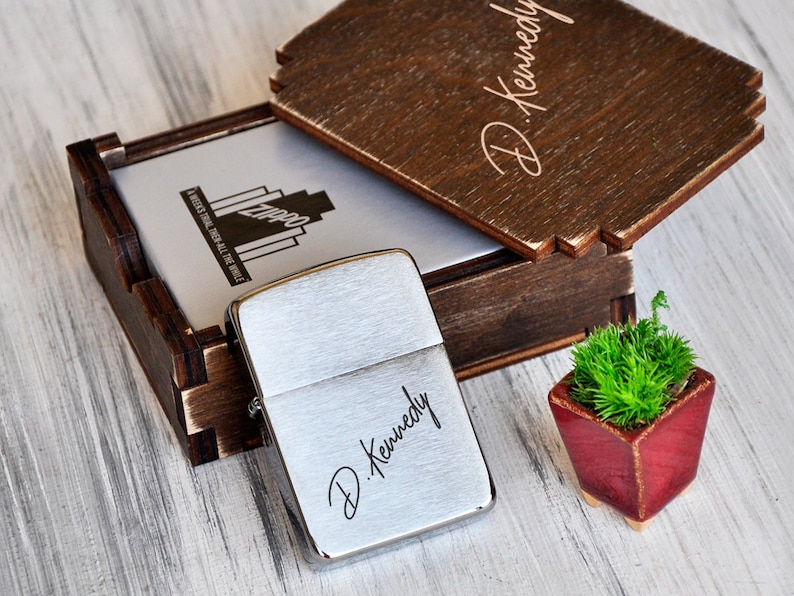 May include: A silver lighter with the name "D. Kennedy" engraved on it. The lighter is in a wooden box with a lid that has the same name engraved on it. The box is sitting on a light wood surface.