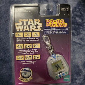 May include: A Star Wars R2-D2 electronic virtual pet toy in its original packaging. The toy is a keychain with a small screen displaying a pixelated image of a character. The packaging features the Star Wars logo and text.