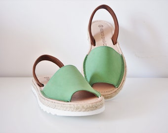 Puede incluir: Un par de sandalias de esparto verdes con correas marrones y suelas blancas. Las sandalias tienen un ribete de cuerda de yute alrededor de la suela.