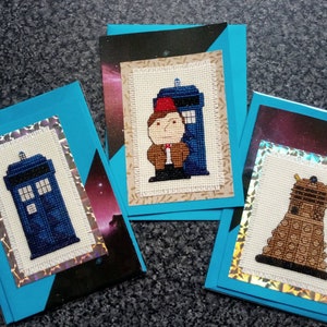 Op de afbeelding: Drie handgemaakte wenskaarten met borduurmotieven van een blauwe politiekast, een bruine Dalek en een man met bruin haar in een bruine jas en rode fez die naast een blauwe politiekast staat. De kaarten hebben een blauwe achtergrond met een zilveren glitterrand.