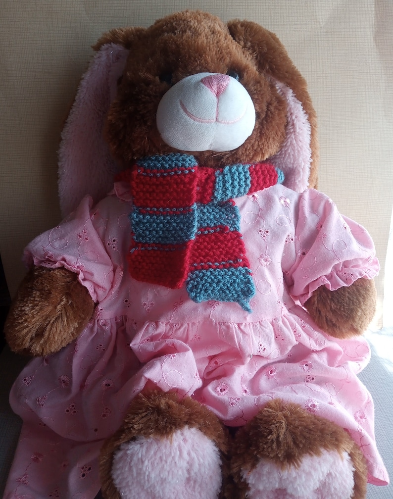 May include: A brown plush bunny wearing a pink dress and a red and blue striped scarf.