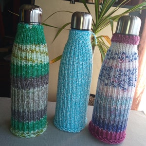 Puede incluir: Tres botellas de agua con cubiertas de punto. Las cubiertas son de diferentes colores y patrones. La botella de la izquierda tiene una cubierta verde, blanca y gris. La botella del medio tiene una cubierta azul claro. La botella de la derecha tiene una cubierta rosa, blanca y azul.
