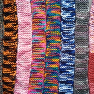 May include: A collection of colorful, knitted scarves. The scarves display a variety of colors, including pink, orange, blue, and gray. Each scarf has a unique pattern and texture, showcasing the craftsmanship of the knitwork.