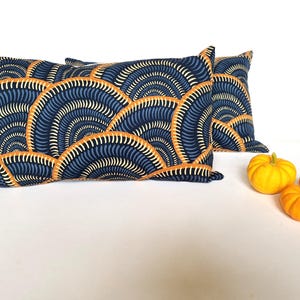 Puede incluir: Dos cojines rectangulares con un diseño en forma de abanico azul marino, naranja y crema. Tres pequeñas calabazas de color amarillo brillante están dispuestas a la derecha de los cojines sobre una superficie blanca.