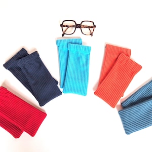 Puede incluir: Una colección de cuatro pares de calentadores de brazos acanalados en rojo, azul oscuro, azul brillante y naranja, dispuestos en semicírculo alrededor de unas gafas con patrón de carey marrón y montura cuadrada grande.