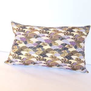 Peut inclure: Un coussin décoratif avec un motif géométrique de vagues superposées de couleur violette, dorée et marron. Le coussin est rectangulaire et a un fond blanc.
