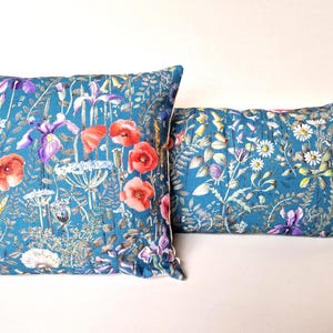 Puede incluir: Dos almohadas de color azul turquesa con un patrón floral que incluye amapolas rojas, iris morados y otras flores silvestres. Las almohadas están hechas de una tela suave y texturizada.