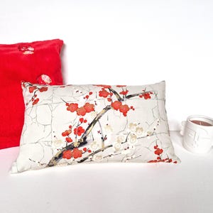 May include: A white pillow with a red and white floral pattern. The pillow has a branch with red flowers and white blossoms. The pillow is on a white surface.