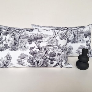 May include: Two rectangular throw pillows featuring a black and white toile de jouy pattern depicting a jungle scene with a leopard, birds, and trees. A small, matte black, stacked-shape decorative vase sits to the right of the pillows.