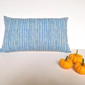 May include: A rectangular pillow with a blue and white striped pattern. The pillow is placed on a white surface, with three small orange pumpkins to the right. The pillow is a decorative item.