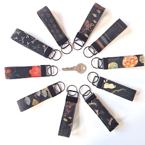 May include: A collection of black fabric keychains with various Japanese-inspired patterns, including dragonflies, fans, and floral designs. Each keychain has a black metal ring and a key in the center. The keychains are arranged in a circular pattern.