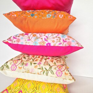 Può includere: Una pila di cinque cuscini decorativi colorati con motivi floreali ricamati e paillettes. I cuscini sono disposti a forma di piramide, con il cuscino più grande in basso e il più piccolo in alto. I cuscini sono tutti realizzati con tessuti diversi e hanno colori diversi, tra cui rosa, arancione, giallo, bianco e blu.