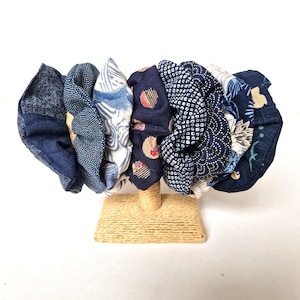 May include: A collection of seven fabric hair scrunchies displayed on a tan rope-wrapped stand. The scrunchies feature various blue and white patterns, including geometric shapes, floral motifs, wave designs, and polka dots in different shades.