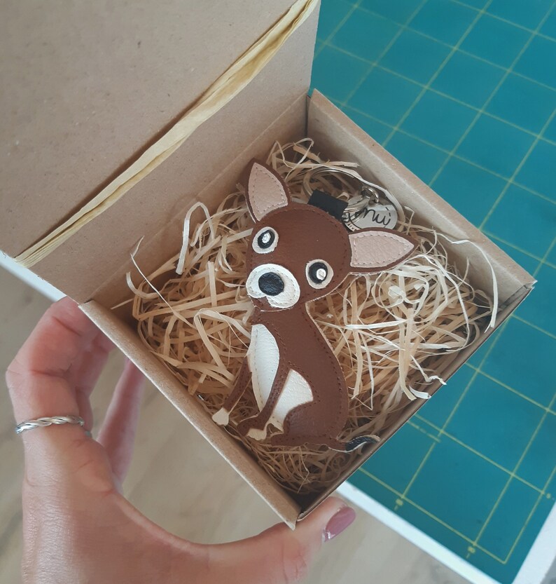 May include: A brown leather chihuahua keychain with white accents, sitting in a box filled with shredded paper. The keychain has a metal ring and a small metal tag with the word "mini" on it.