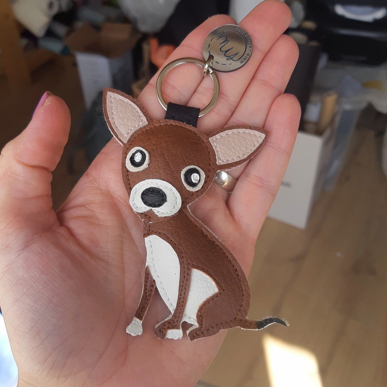 May include: Brown leather chihuahua keychain with white accents and black eyes. The keychain has a silver ring and a silver tag that says "Mus" on it.