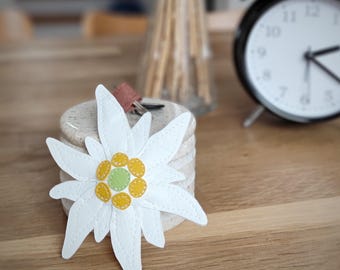 May include: A white leather edelweiss flower keychain with yellow and green stitching. The keychain is sitting on a marble coaster.
