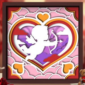 May include: A layered paper art piece featuring a white Cupid aiming a bow and arrow within a heart-shaped design. The artwork uses shades of pink, red, orange, and purple, set in a brown frame. The theme is love and romance.
