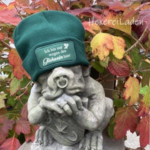 Peut inclure: Un bonnet vert foncé avec un patch rectangulaire portant l'inscription "Ich bin nur wegen des Glühwein hier" en lettres blanches. Le bonnet est posé sur une statue de gargouille en pierre. Le fond présente des feuilles d'automne colorées.