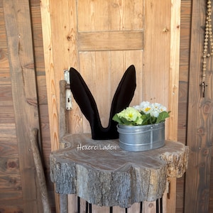 Può includere: Orecchie di coniglio in velluto nero sedute su un ceppo di legno con un secchio di metallo pieno di fiori bianchi e gialli.