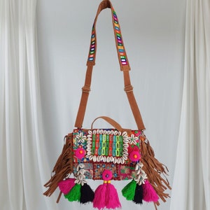 May include: A colorful patchwork crossbody bag with a brown leather strap, fringe trim, and shell accents. The bag has a variety of colorful embroidery and tassels.
