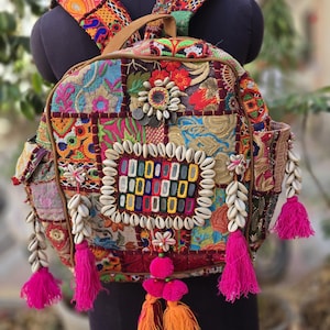 Op de afbeelding: Een rugzak van veelkleurige patchworkstof met ingewikkeld borduurwerk, kaurischelpversieringen en felroze en oranje kwastjes. De tas heeft een centraal vierkant patroon, verstelbare schouderbanden en zijvakken.