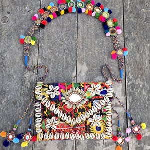 May include: A colorful, embroidered crossbody bag with a matching headband. The bag features a black background with floral and geometric patterns, adorned with seashells and a central mirror. The headband is decorated with mirrors and colorful pom-poms.