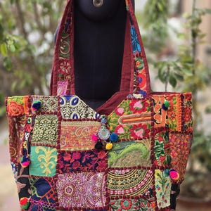 May include: A large tote bag constructed from a patchwork of multicolored embroidered fabrics featuring floral and geometric patterns. The bag includes a wide shoulder strap, decorative hanging pom-poms, and a central circular mirror accent.
