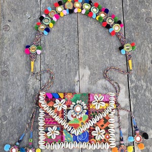 May include: A colorful, bohemian-style handbag and matching headband. The bag features a patchwork design with white seashell trim and floral accents. The headband is adorned with colorful pom-poms and mirrored embellishments. Both items have intricate details and a vibrant aesthetic.