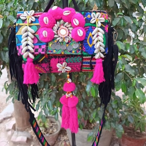 Può includere: Una colorata borsa ricamata con frange e cinturino in camoscio nero. La borsa presenta pompon rosa, conchiglie bianche e monete decorative. Il cinturino è decorato con un motivo arcobaleno.