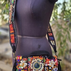 May include: A black crossbody bag with a colorful embroidered design, adorned with seashells and a central mirror. The bag features a blue tassel fringe and a patterned strap, showcasing a bohemian aesthetic.