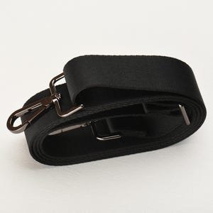 Puede incluir: Una correa de hombro negra ajustable con herrajes de metal plateado. La correa está hecha de un material tejido duradero y cuenta con un gancho giratorio y una hebilla deslizante para ajustar la longitud. Ideal para bolsos u otros accesorios.