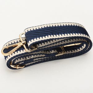 May include: A navy blue and cream-coloured woven bag strap with gold-coloured metal hardware. The strap features a repeating geometric pattern and a gold-coloured clasp for easy attachment to a bag. The strap is approximately 3.8 cm wide.