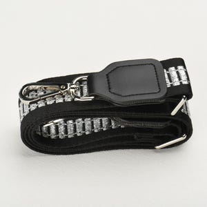 以下が含まれることがあります： 黒とシルバーのバッグストラップ。シルバーの留め金と黒革のアクセントが付いています。ストラップは、黒い背景に繰り返されるシルバーのパターンが特徴です。ストラップは調整可能で、バッグに取り付けることができます。