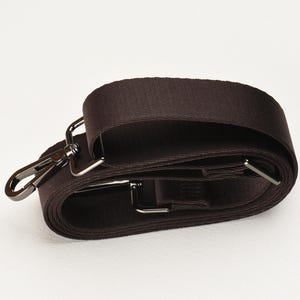 May include: A dark brown bag strap with silver-coloured metal hardware. The strap is folded over itself, showing the texture of the material. The metal hardware includes a swivel clasp and rectangular loops.