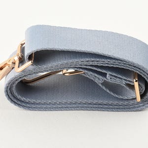 Puede incluir: Una correa de bolso azul claro con herrajes metálicos dorados. La correa está hecha de tela tejida y tiene un cierre giratorio y hebillas ajustables. La correa está enrollada, mostrando su textura y construcción.