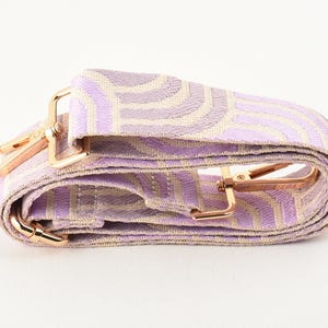 May include: A folded bag strap with a repeating wave pattern in lavender and beige. It has gold-coloured metal clasps for attachment. The strap is designed to be attached to a handbag or shoulder bag.