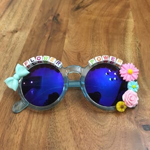 May include: Blue round sunglasses with a flower power theme. The sunglasses have blue lenses and are decorated with colorful plastic flowers and letter beads that spell out "flower power".